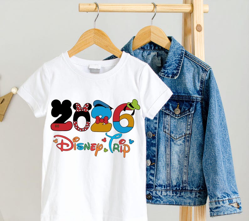 Op de afbeelding: Wit T-shirt met een kleurrijke "2026 Disney Trip" afbeelding. De cijfers zijn ontworpen met Disney-karakterelementen. Een spijkerjasje hangt achter het T-shirt aan een houten rek.
