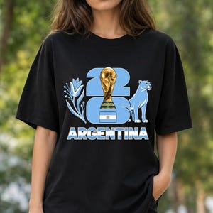 Puede incluir: Camiseta negra con un dise&ntilde;o gr&aacute;fico que celebra la victoria futbol&iacute;stica de Argentina. El dise&ntilde;o incluye un trofeo dorado, un puma azul y la palabra "ARGENTINA" en letras blancas. Tambi&eacute;n aparece el n&uacute;mero "23".