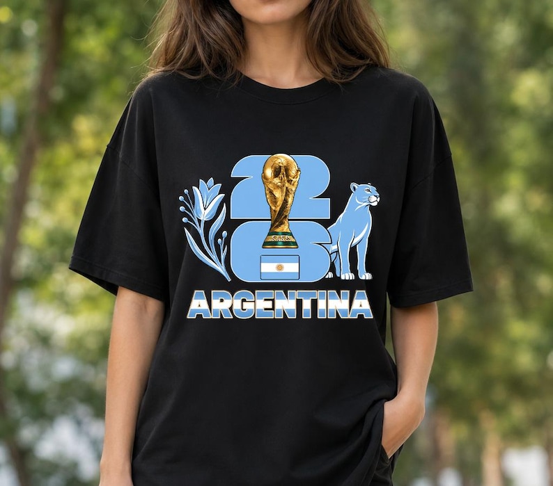 Puede incluir: Camiseta negra con un dise&ntilde;o gr&aacute;fico que celebra la victoria futbol&iacute;stica de Argentina. El dise&ntilde;o incluye un trofeo dorado, un puma azul y la palabra "ARGENTINA" en letras blancas. Tambi&eacute;n aparece el n&uacute;mero "23".