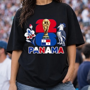 Puede incluir: Camiseta negra con un dise&ntilde;o gr&aacute;fico que presenta el n&uacute;mero "22", un trofeo dorado, una flor azul y blanca, un &aacute;guila estilizada y la palabra "PANAM&Aacute;" en rojo y blanco. La camiseta tiene mangas cortas.