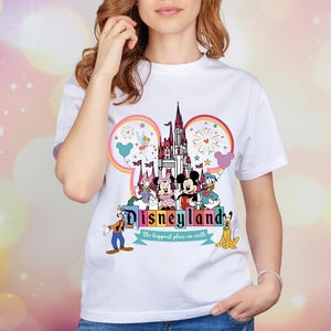 Op de afbeelding: Wit T-shirt met een kleurrijke Disneyland-afbeelding. Het ontwerp toont het Disneyland-logo, een kasteel en Disney-personages zoals Mickey en Minnie Mouse, Donald Duck en Goofy. De woorden "Disneyland" en "the happiest place on earth" zijn ook aanwezig.