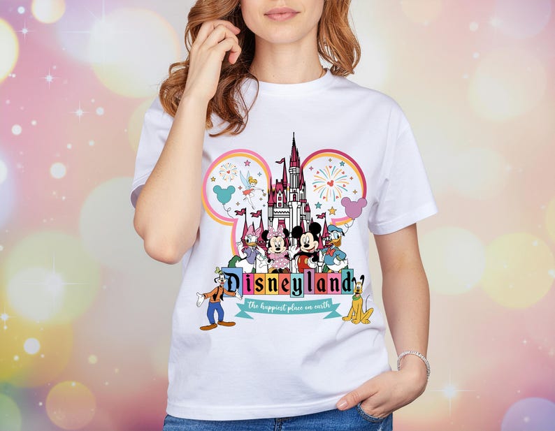 Op de afbeelding: Wit T-shirt met een kleurrijke Disneyland-afbeelding. Het ontwerp toont het Disneyland-logo, een kasteel en Disney-personages zoals Mickey en Minnie Mouse, Donald Duck en Goofy. De woorden "Disneyland" en "the happiest place on earth" zijn ook aanwezig.