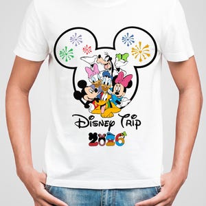 Op de afbeelding: Wit T-shirt met een kleurrijke afbeelding van Disney-personages, waaronder Mickey en Minnie Mouse, Donald Duck en Goofy. Het ontwerp is omlijst door een Mickey Mouse-hoofdvorm en bevat de tekst "Disney Trip 2026".