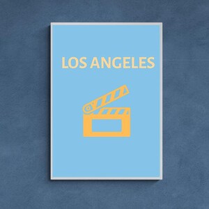 Puede incluir: Un póster enmarcado con fondo azul claro. Las palabras "LOS ANGELES" están impresas en color naranja claro en la parte superior. Debajo, un icono de claqueta de cine naranja claro.