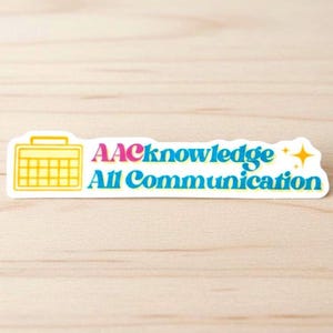 May include: A rectangular sticker with the text "AACknowledge All Communication" in blue, pink, and white. A yellow calendar icon is on the left, and a yellow star is on the right. The sticker is on a light wood surface.