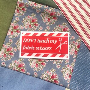 May include: A patchwork textile with a red sticker that says "DON'T touch my fabric scissors." The sticker has white diagonal stripes and text. The fabric has floral patterns in pink and red on a blue background.