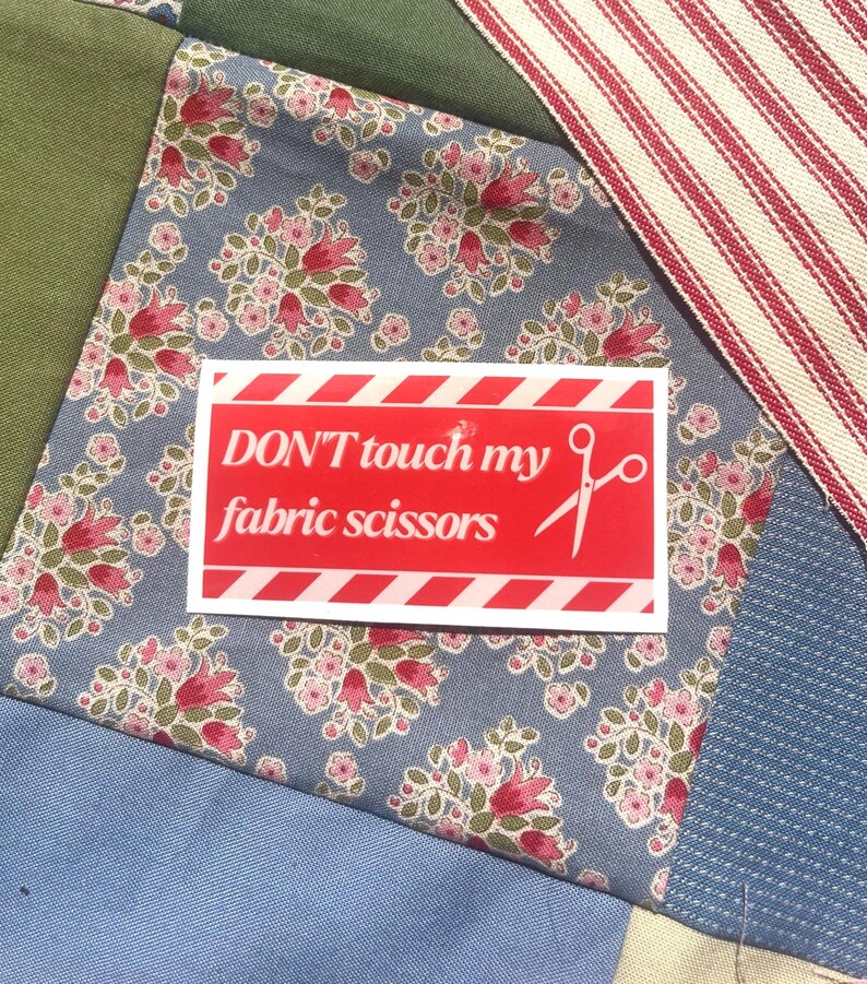 May include: A patchwork textile with a red sticker that says "DON'T touch my fabric scissors." The sticker has white diagonal stripes and text. The fabric has floral patterns in pink and red on a blue background.