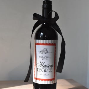 May include: A dark wine bottle with a black bow. The label reads "Pairs Well With Master's Degree" in a decorative font, with a graduation cap illustration. The bottle is a celebratory gift for a graduate.