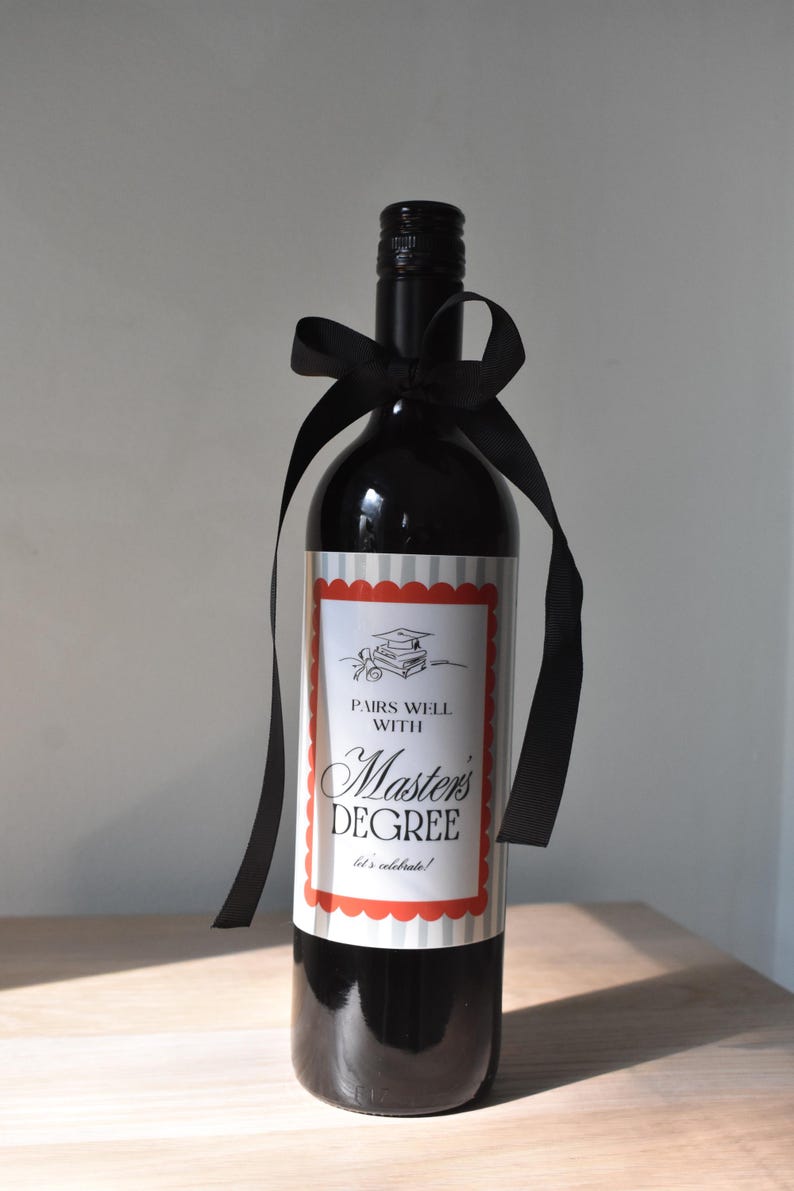 May include: A dark wine bottle with a black bow. The label reads "Pairs Well With Master's Degree" in a decorative font, with a graduation cap illustration. The bottle is a celebratory gift for a graduate.