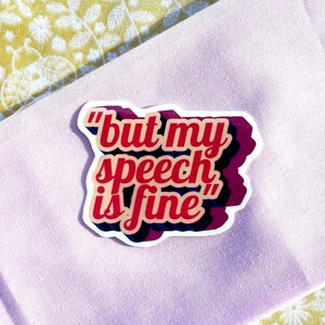 May include: A sticker with the phrase "but my speech is fine" in red, pink, and black lettering. The sticker is on a light pink fabric, with a yellow floral patterned fabric in the background.