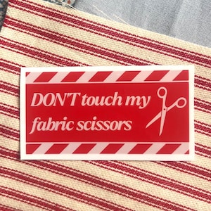 May include: A red sticker with white diagonal stripes and the text "DON'T touch my fabric scissors" in white. A white scissors graphic is on the right. The sticker is on a red and white striped fabric.