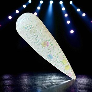 May include: A clear, elongated nail with iridescent glitter and colorful circular accents. The nail is set against a dark background with stage lighting, creating a glamorous effect. The nail is a light, translucent color.