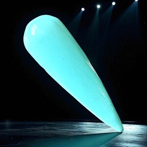 May include: A single, elongated nail tip in a vibrant, glowing teal color. The nail is smooth and glossy, with a pointed shape. The background is dark, with spotlights illuminating the nail, creating a dramatic effect. The nail is standing upright.