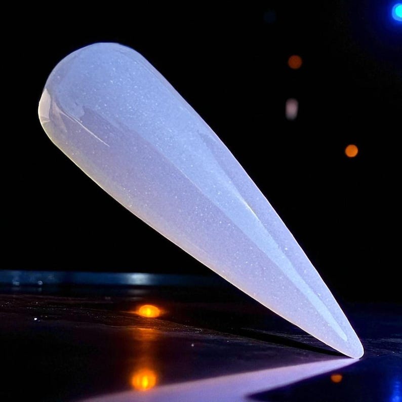 May include: A light blue, elongated nail with a pointed tip. The nail has a pearlescent, glittery finish. The background is dark, with a few small, blurred lights.