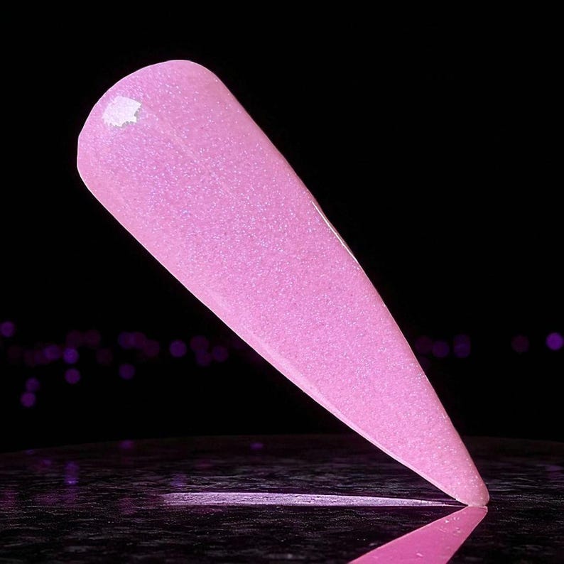 May include: A single, light pink nail with a glittery finish. The nail is long and pointed, with a smooth, glossy surface. The background is black, with a blurred reflection of the nail.