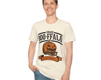 May include: Cream-colored t-shirt with a graphic featuring a jack-o'-lantern wearing a football helmet. The text reads "1960 BOO-FFALO FOOTBALL" in black and orange. The shirt is worn by a person in blue jeans.