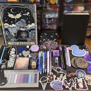 May include: A collection of stationery and accessories, including a large zippered pouch with "THE READER" text, a black notebook, pens, stickers, and decorative items. The color palette is primarily purple, black, and blue, with a celestial and gothic theme.
