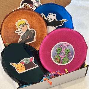 May include: A box containing four colorful, round coasters. The coasters are orange, blue, pink, and dark green, each with a different sticker design. The stickers feature a panda, aliens, and cartoon characters.