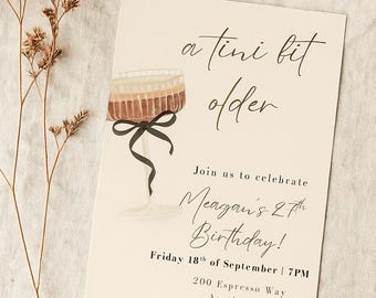 May include: A cream-colored birthday invitation with the text "a tini bit older" and "Meagan's 27th Birthday!" A watercolor illustration of a cocktail glass with a black ribbon is featured. Dried floral stems are in the background.