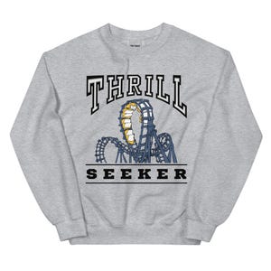 Könnte beinhalten: Ein hellgraues Sweatshirt mit einem Achterbahn-Motiv. Das Wort "THRILL" ist in schwarzen und weißen Blockbuchstaben oben aufgebogen. Unter der Achterbahn steht das Wort "SEEKER" in schwarzen Blockbuchstaben.