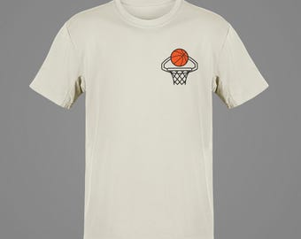 May include: Cream-colored t-shirt featuring a basketball graphic. The design includes a basketball and a hoop, rendered in orange and black. The shirt has short sleeves and a crew neck.