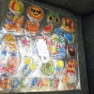 May include: A sheet of colorful summer-themed stickers. Designs include a beach ball, sunglasses-wearing sun, watermelon slice, crab, and the words "Hello Summer" and "Pool Party".