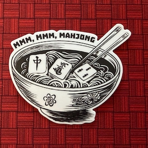 May include: A black and white sticker featuring a bowl of noodles with Mahjong tiles and chopsticks. The text "MMM, MMM, MAHJONG" is above the bowl. The sticker is on a red checkered background.