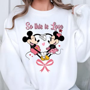 Pu&ograve; includere: Felpa bianca con un disegno di Topolino e Minnie che formano un cuore con le mani. Il design include cuori rosa, un fiocco rosa e la scritta "So this is Love" in rosa. I personaggi dei cartoni animati indossano i loro abiti classici.