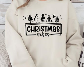 May include: A cream-colored sweatshirt with a black graphic of stylized Christmas trees and the words "CHRISTMAS vibes" in a rectangular frame. The design includes stars and a simple font.