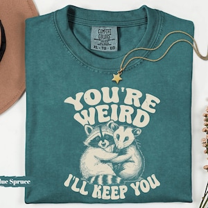 May include: Blue Spruce t-shirt with the text "You're Weird, I'll Keep You" and a graphic of two hugging raccoons. A gold star pendant necklace rests on the shirt. The shirt has a Comfort Colors tag, size XL-TG-EG.