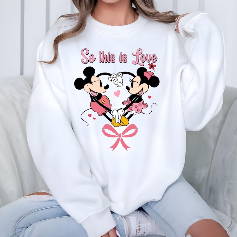 Pu&ograve; includere: Felpa bianca con un disegno di Topolino e Minnie che formano un cuore con le mani. Il design include cuori rosa, un fiocco rosa e la scritta "So this is Love" in rosa. I personaggi dei cartoni animati indossano i loro abiti classici.