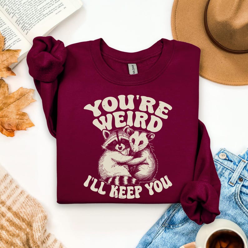May include: Burgundy crewneck sweatshirt with the text "YOU'RE WEIRD I'LL KEEP YOU" in white. The graphic features a raccoon and opossum hugging. The sweatshirt is made of a soft material.