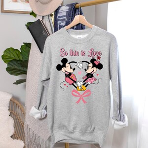 Pu&ograve; includere: Una felpa grigio melange con una grafica di Topolino e Minni che formano un cuore con le mani. Il testo "So this is Love" &egrave; sopra la grafica in rosa. Un fiocco rosa &egrave; nella parte inferiore della grafica.