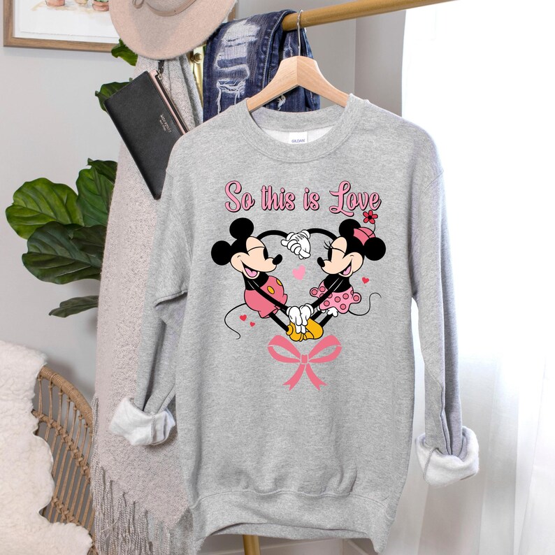 Pu&ograve; includere: Una felpa grigio melange con una grafica di Topolino e Minni che formano un cuore con le mani. Il testo "So this is Love" &egrave; sopra la grafica in rosa. Un fiocco rosa &egrave; nella parte inferiore della grafica.