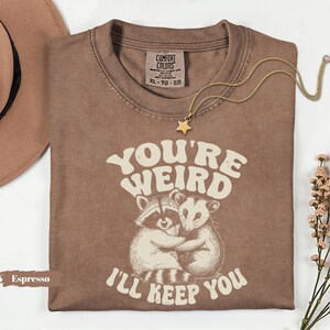 May include: Espresso-colored t-shirt with the text "YOU'RE WEIRD I'LL KEEP YOU" and an illustration of two hugging animals. A gold star pendant necklace and a brown hat are also visible.