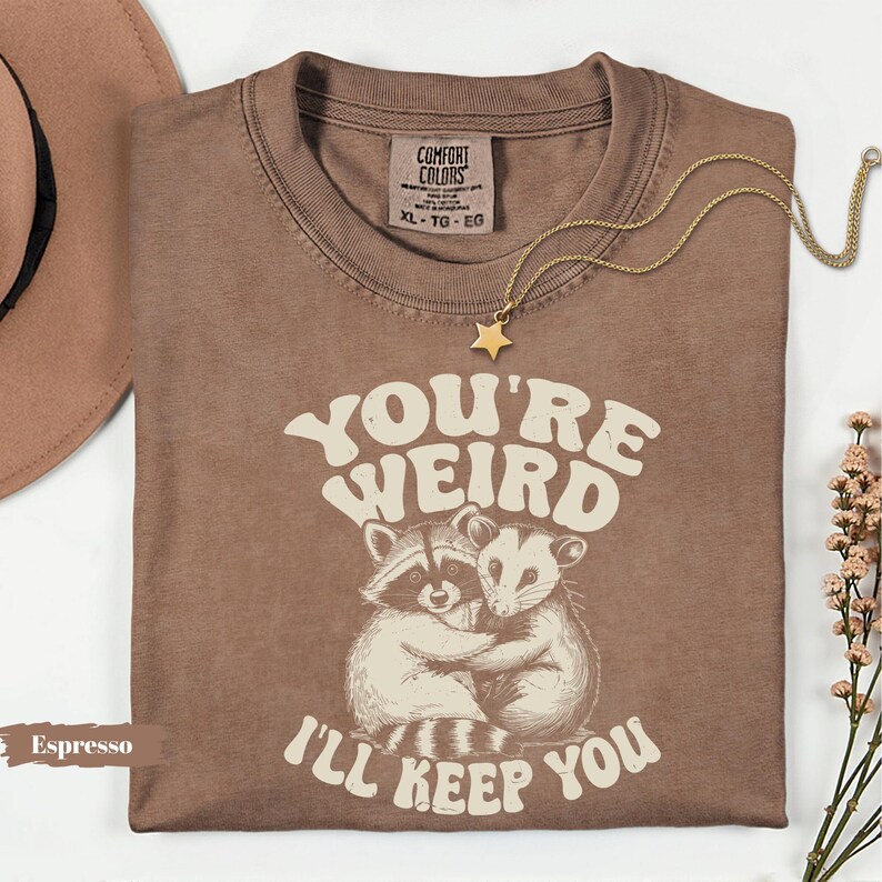 May include: Espresso-colored t-shirt with the text "YOU'RE WEIRD I'LL KEEP YOU" and an illustration of two hugging animals. A gold star pendant necklace and a brown hat are also visible.