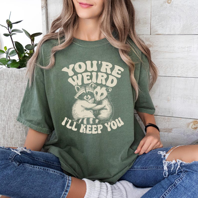 May include: Olive green t-shirt with the text "YOU'RE WEIRD I'LL KEEP YOU" above an illustration of two hugging raccoons. The shirt has a vintage, slightly faded look.