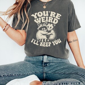 May include: Dark gray t-shirt with the text "YOU'RE WEIRD I'LL KEEP YOU" in a retro font. The shirt features a graphic of a raccoon and opossum hugging. The shirt is made of a soft material and has a vintage look.