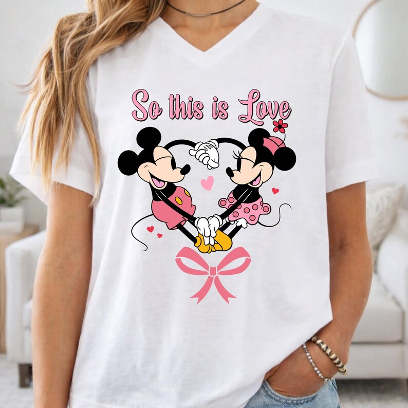 May include: White V-neck t-shirt featuring a graphic of Mickey and Minnie Mouse forming a heart with their hands. The text "So this is Love" is above the characters, with a pink bow at the bottom.