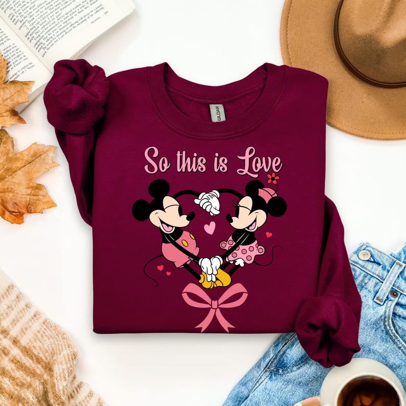 Pu&ograve; includere: Felpa bordeaux con un design a cuore di Minnie e Topolino e la scritta "So this is Love". La grafica include cuori rosa e un fiocco. La felpa &egrave; abbinata a jeans blu e un cappello marrone.