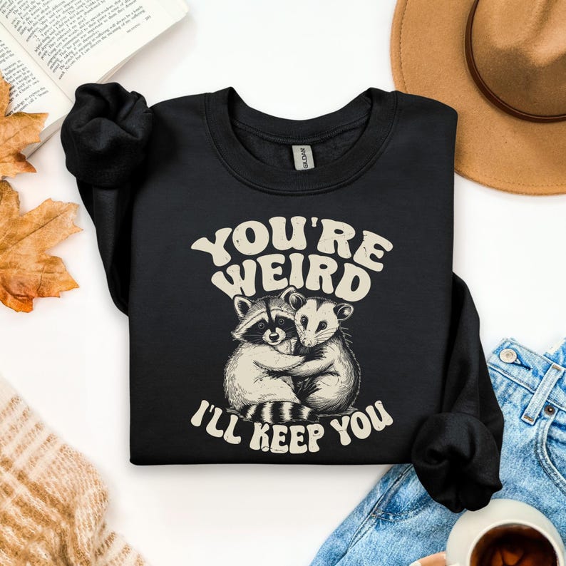 May include: Black sweatshirt with the text "YOU'RE WEIRD I'LL KEEP YOU" in a retro font. The graphic features a raccoon and opossum hugging. The sweatshirt is a crew neck style.
