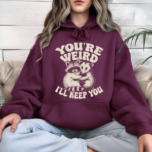 May include: A maroon hoodie with a drawstring hood and a kangaroo pocket. The front features a graphic of a raccoon and opossum hugging, with the text "YOU'RE WEIRD I'LL KEEP YOU" in a retro font.