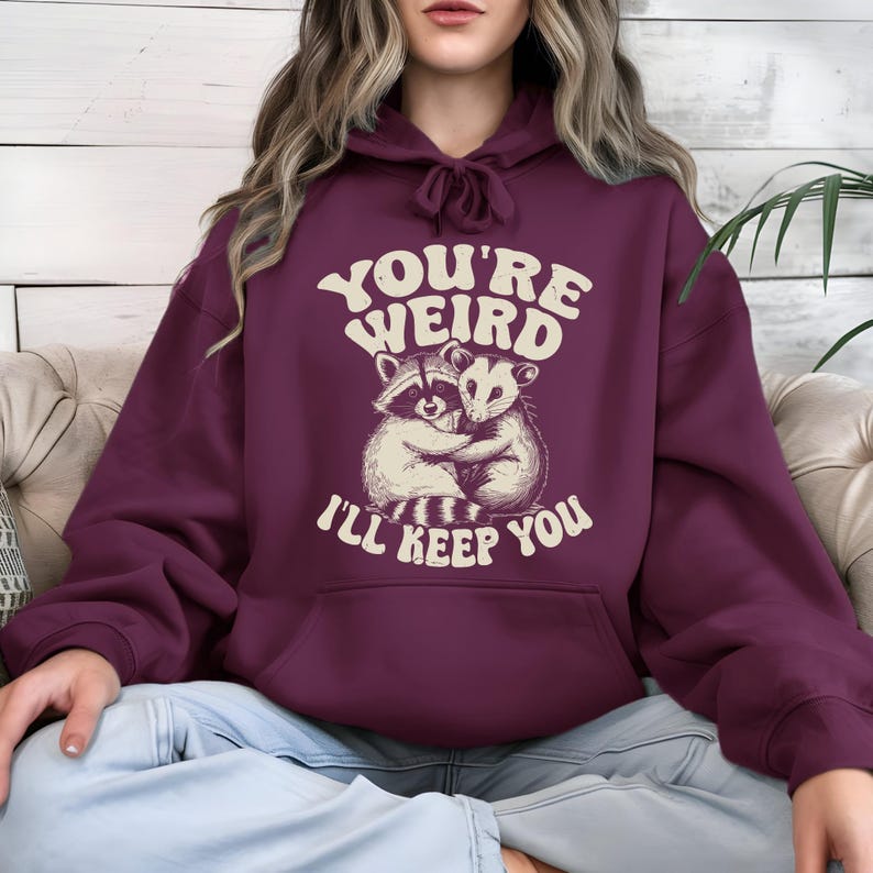 May include: A maroon hoodie with a drawstring hood and a kangaroo pocket. The front features a graphic of a raccoon and opossum hugging, with the text "YOU'RE WEIRD I'LL KEEP YOU" in a retro font.