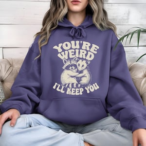 May include: A periwinkle blue hoodie with a drawstring hood and a kangaroo pocket. The front features a graphic of a raccoon and opossum hugging, with the text "YOU'RE WEIRD I'LL KEEP YOU" in a retro font.