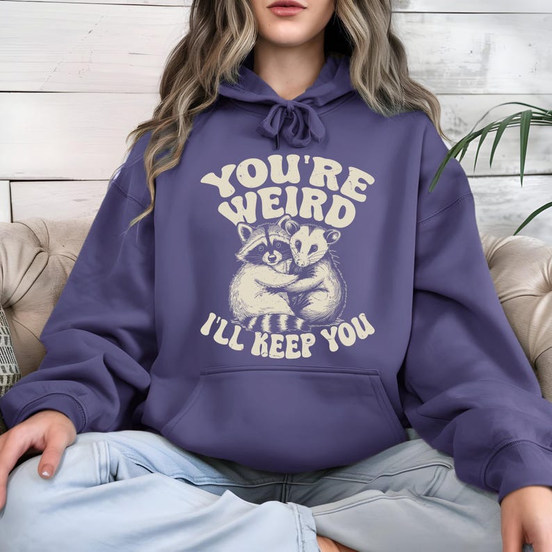 May include: A periwinkle blue hoodie with a drawstring hood and a kangaroo pocket. The front features a graphic of a raccoon and opossum hugging, with the text "YOU'RE WEIRD I'LL KEEP YOU" in a retro font.