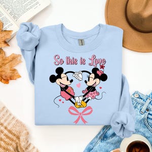 Pu&ograve; includere: Felpa azzurra con Topolino e Minnie che si tengono per mano a forma di cuore. Il testo "So this is Love" &egrave; sopra i personaggi. Minnie indossa un vestito rosa a pois e Topolino indossa pantaloncini rosa. Un fiocco rosa &egrave; in basso.