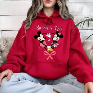 Pu&ograve; includere: Felpa con cappuccio rossa con un disegno di Topolino e Minnie che formano un cuore con le mani. Il testo "So this is Love" &egrave; sopra il disegno. Un fiocco rosa &egrave; nella parte inferiore del cuore.