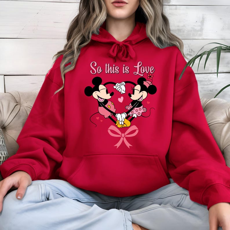Pu&ograve; includere: Felpa con cappuccio rossa con un disegno di Topolino e Minnie che formano un cuore con le mani. Il testo "So this is Love" &egrave; sopra il disegno. Un fiocco rosa &egrave; nella parte inferiore del cuore.