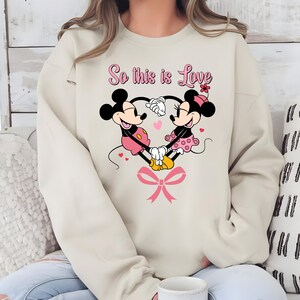 Pu&ograve; includere: Felpa beige con un design a forma di cuore con Topolino e Minni che si tengono per mano. La frase "So this is Love" &egrave; scritta sopra i personaggi in rosa, con un fiocco rosa in basso.