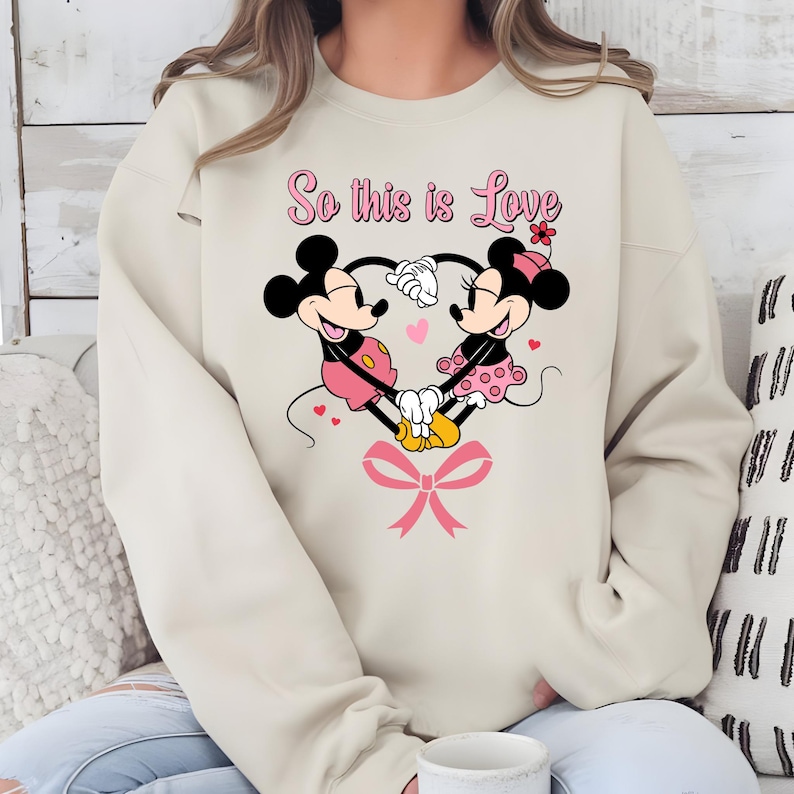 Pu&ograve; includere: Felpa beige con un design a forma di cuore con Topolino e Minni che si tengono per mano. La frase "So this is Love" &egrave; scritta sopra i personaggi in rosa, con un fiocco rosa in basso.